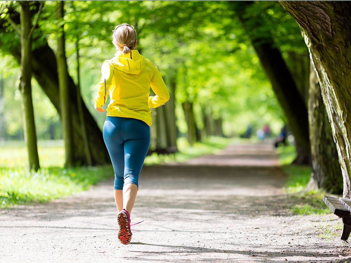 Kobieta uprawiająca jogging po leśnej ścieżce, ubrana w żółtą kurtkę i niebieskie legginsy. - zdjęcie